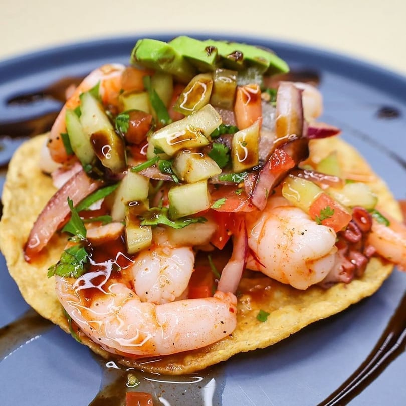 Shrimp and octopus ceviche tostada topped with avocado, cucumber, and a dark sauce.