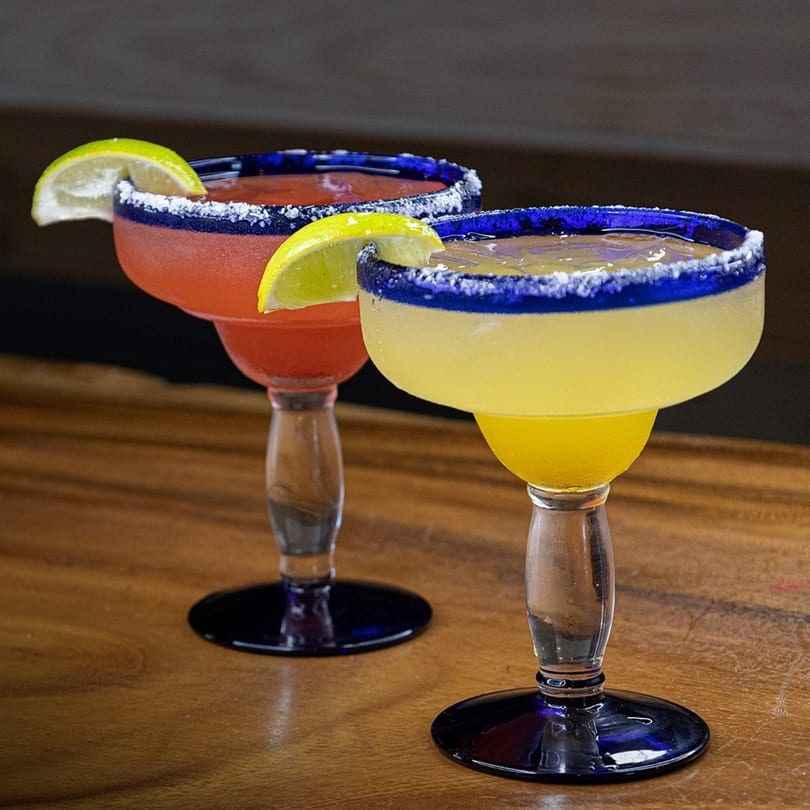 Two vibrant margaritas, one red and one yellow, with salted rims and lime wedges on a wooden table.