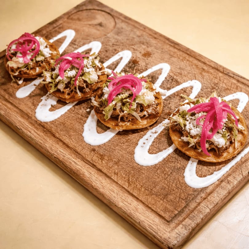 Four sopes with shredded meat, lettuce, cheese, and pickled red onions on a wooden board.